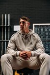Man in beige trainingsjas en broek, zittend in een sportschool.