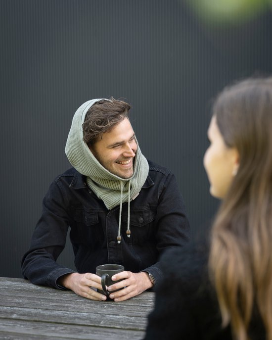 Een man met een grijze spijkerjas en een lichtgrijze gebreide colsjaal met capuchon die hij aantrekt.