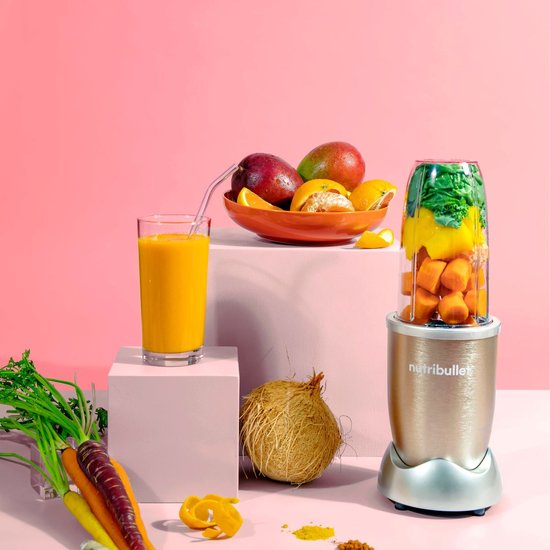 Nutribullet blender in champagnekleur op een roze display, naast een glas smoothie en diverse vruchten.