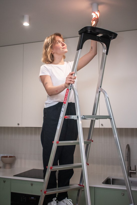 Een vrouw staat op een aluminium Baulich trap in een keuken, een lamp aan het plafond vervangend.