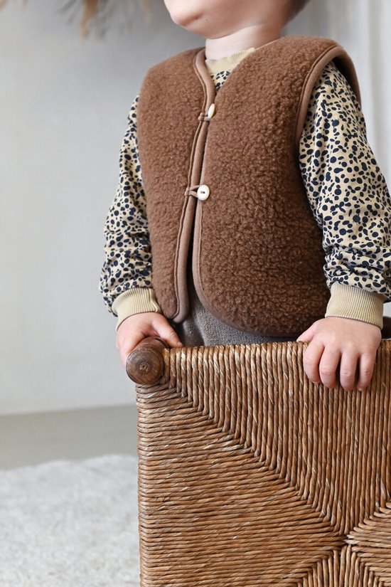 Kind draagt bruin wollen vest met bruine biezen en twee knopen over een shirt met luipaardprint.