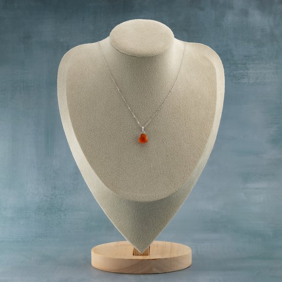 Zilveren ketting met een kleine, hartvormige oranje carneool hanger, gedrapeerd om een beige buste.