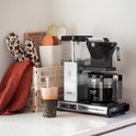Zilverkleurige Moccamaster Select koffiezetapparaat met glazen kan, naast accessoires zoals potjes en handdoeken.