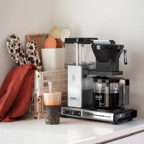 Zilverkleurige Moccamaster Select koffiezetapparaat met glazen kan, naast accessoires zoals potjes en handdoeken.