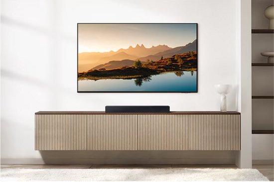 Zwarte soundbar onder een tv waarop een landschap met bergtoppen, een meer en bomen wordt getoond.