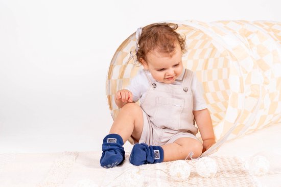 Een baby zit in een speeltunnel, met donkerblauwe fleece Little Riots slofjes aan.