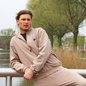 Man draagt een taupekleurige sweater met ritssluiting, logo op de borst, bij een reling.