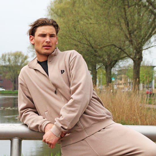 Man draagt een taupekleurige sweater met ritssluiting, logo op de borst, bij een reling.