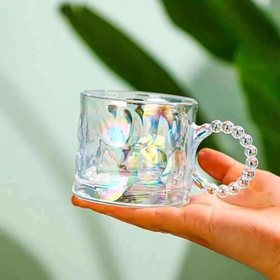 Tasse en verre avec un effet nacré, posée dans une main. L'anse est ornée de perles en verre.