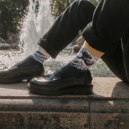 Een persoon zittend bij een fontein draagt donkerblauwe sokken met de print van 'De Grote Golf van Kanagawa' en zwarte loafers.