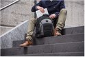 Man in donkere jas en chino's zit op trap en pakt laptop uit zwarte rugzak met grijze en rode details.