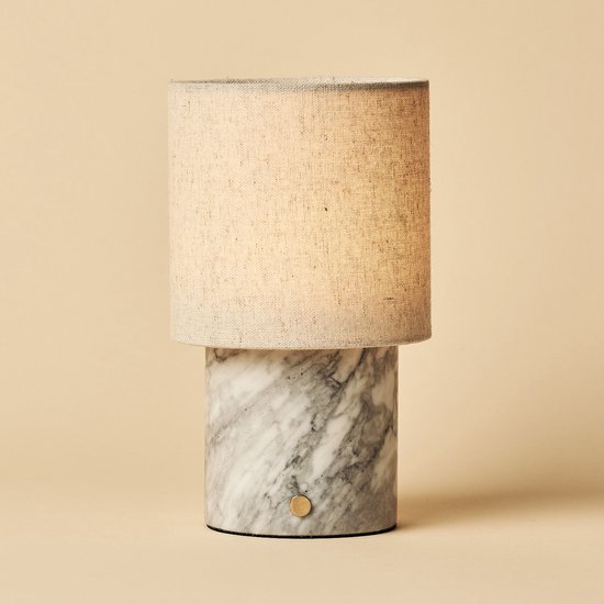 Een marmeren tafellamp met een beige linnen kap, met gouden knop, op een beige achtergrond.