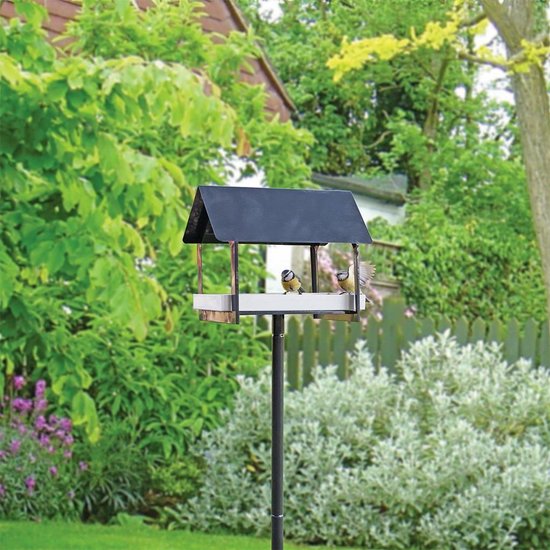 Zwarte voederhuis op paal met witte voerbak met zaadjes, twee mezen eten ervan in een tuin.