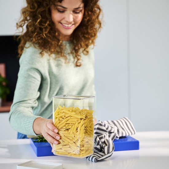 Une femme souriante aux cheveux bouclés tient une boîte de conservation transparente remplie de pâtes penne. Un torchon rayé et une planche à découper bleue sont visibles.