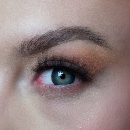 Close-up van het oog van een vrouw met een blauwgroene contactlens, met bruine oogschaduw en lange wimpers.