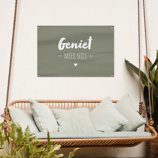 Donkergroene banner met witte tekst 'Geniet - Meer Niet -' en een hartje, hangend aan een witte muur boven een rieten schommelbank.