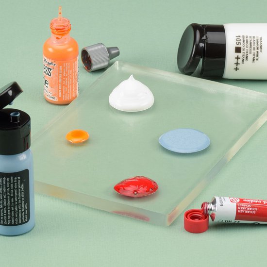 Een transparante gelplaat met druppels witte, oranje, blauwe en rode verf. Naast de plaat staan flesjes oranje en blauwe inkt, een tube witte verf en een tube rode verf.