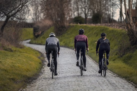 Drie fietsers rijden op een landweg met kasseien. De middelste fietser draagt een bordeauxrood shirt met lange mouwen.