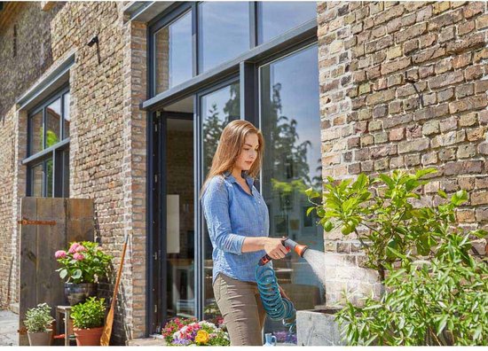 Vrouw in blauw hemd en bruine broek besproeit planten met blauwe spiraalslang bij een bakstenen huis.