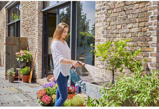 Vrouw in wit hemd en jeans besproeit planten met blauwe spiraalslang bij een bakstenen gevel.