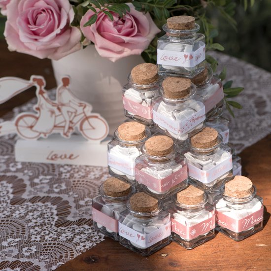 Empilement de mini bouteilles carrées en verre avec bouchon en liège, remplies de bonbons blancs, décorées de rubans avec 'Love' et 'Merci'.