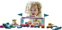 Une jeune fille aux cheveux bouclés regarde une piste de course LEGO Duplo avec trois voitures et des drapeaux.