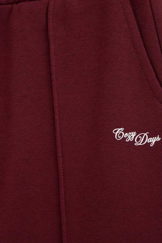 Bordeauxrode joggingbroek met 'Cozy Days' in sierlijk schrift op de zijkant.