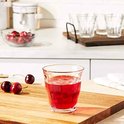 Un verre Duralex Picardie transparent avec cannelures verticales, rempli d'une boisson rouge, sur une planche en bois avec des cerises.
