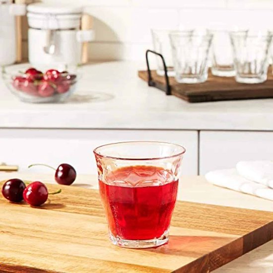 Un verre Duralex Picardie transparent avec cannelures verticales, rempli d'une boisson rouge, sur une planche en bois avec des cerises.