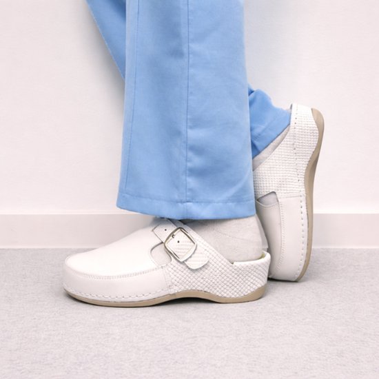 Witte medische klompen met gesp, gedragen met lichtblauwe broek.
