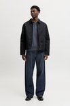 Man draagt een zwarte jas over een donkerblauwe polo en jeans.