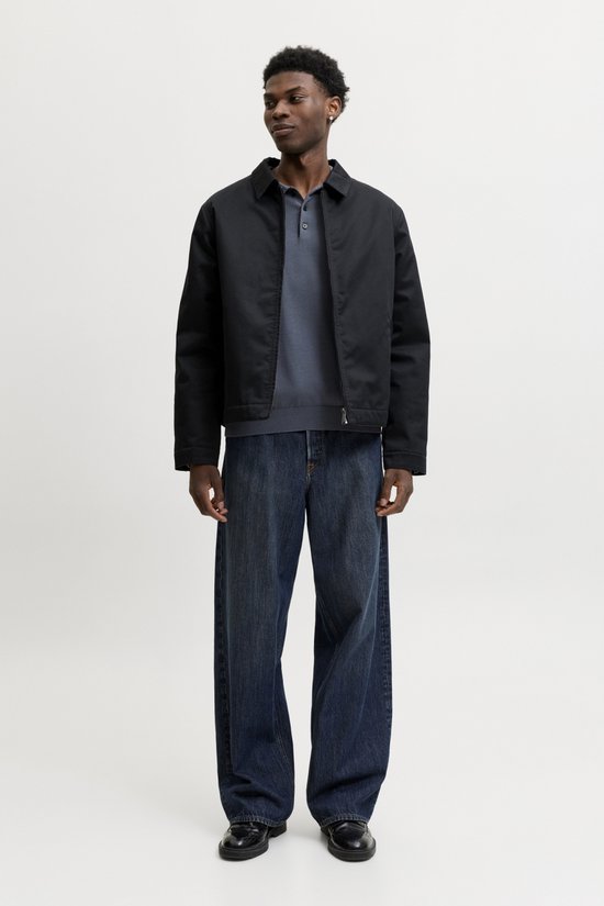Man draagt een zwarte jas over een donkerblauwe polo en jeans.
