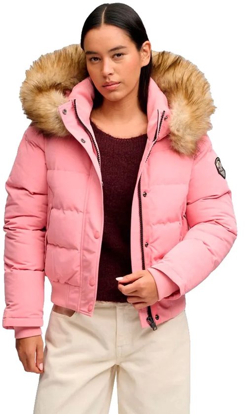 Een jongedame draagt een roze bomberjack met bruine bontkraag, een bordeauxrode coltrui en een beige broek.