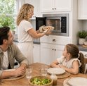 Een gezin aan tafel in de keuken. Een vrouw haalt een schaal met eten uit de Heinner inbouwmagnetron.
