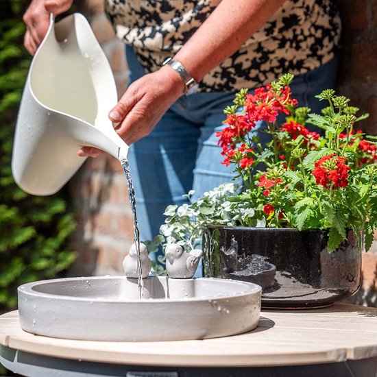 Iemand vult een grijze vogeldrinkschaal met een witte gieter naast rode bloemen in een pot.