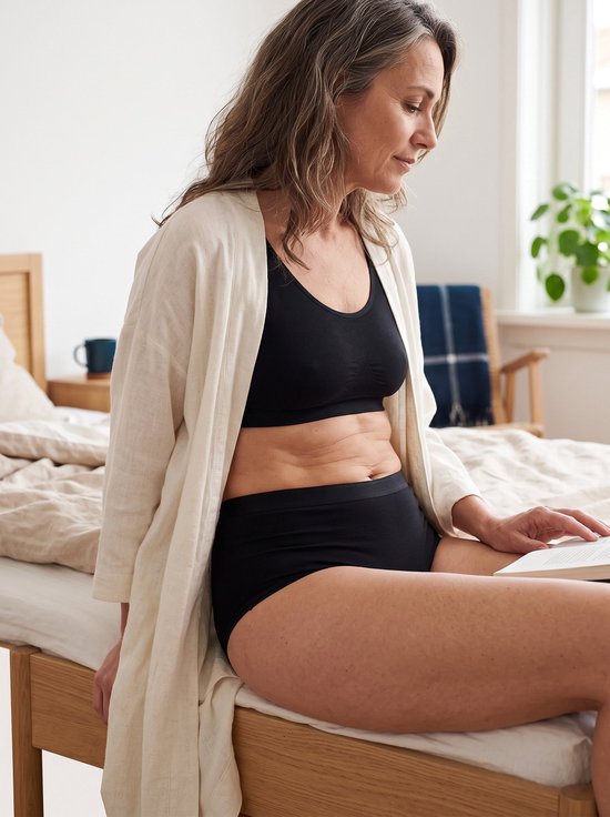 Vrouw draagt zwarte high-waist slip en bh onder een licht beige vest terwijl ze op een bed zit en leest.
