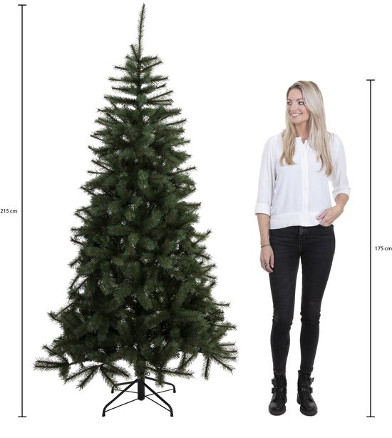 Groene kunstkerstboom van 215 cm naast een vrouw van 175 cm voor de schaalvergroting.
