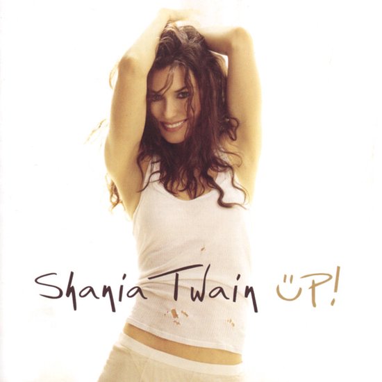 Shania Twain glimlacht met haar handen in haar haar, draagt een wit hemdje met tekst "Up!" op een lichte achtergrond.
