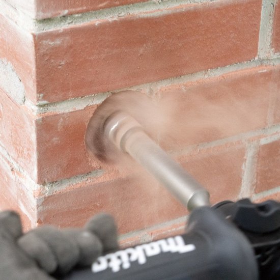 Een Makita-machine boort met een gatzaag in een rode bakstenen muur.