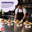 Chef in zwarte schort schikt hamburgers en bruschetta op borden met gouden rand. Tekst: catering, ideaal voor evenementen.
