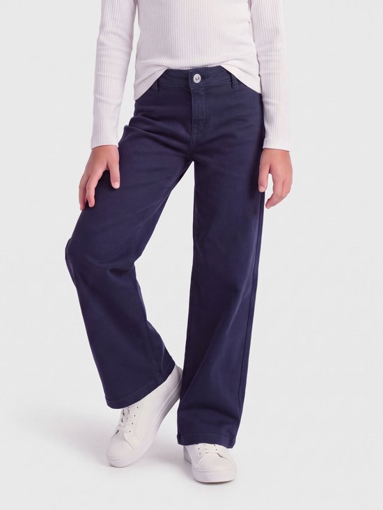 Model draagt marineblauwe jeans met wijde pijpen en witte sneakers, gecombineerd met een witte top met lange mouwen.