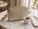 Gouden crossbodytas met twee ritsen en een beige geweven schouderband op een marmeren tafel naast een kopje koffie.
