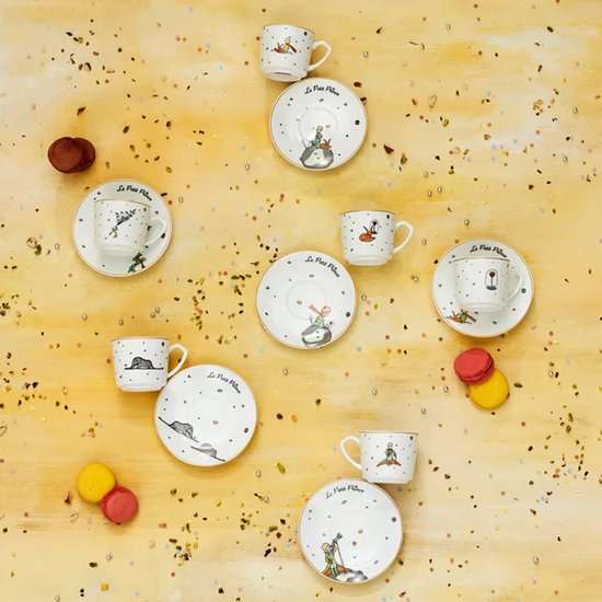 Zes porseleinen espressokopjes en schotels met Le Petit Prince-motieven, verspreid op een gele achtergrond.