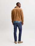 Man in een spijkerbroek, een blauw overhemd en een bruine jas, van achteren gezien.