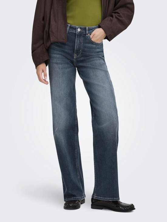 Model in een olijfgroene coltrui en wijde spijkerbroek met een stonewash-look. Een bruine jas wordt vastgehouden.