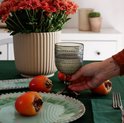 Een hand pakt een donkergrijs glas met bobbelig reliëf, op een tafel met perziken en bloemen.