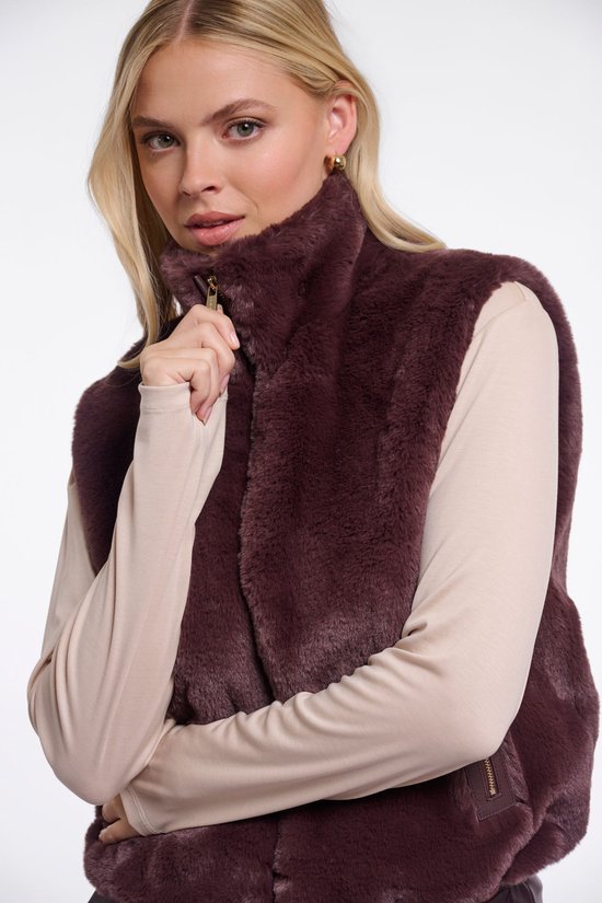 Close-up van een bruin kort gilet met rits en hoge kraag, gedragen door een model met een beige coltrui.