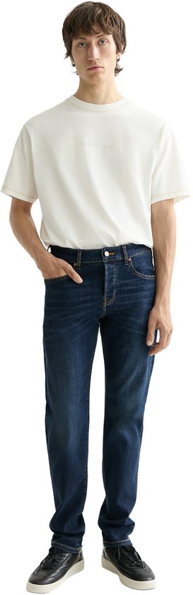 Man in een donkerblauwe jeans en een wit T-shirt. Hij staat met zijn hand in de zak van de jeans.