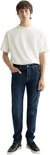 Man in een donkerblauwe jeans en een wit T-shirt. Hij staat met zijn hand in de zak van de jeans.