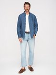 Man met baard staat, draagt een blauw overhemd met witte stipjes, wit t-shirt, spijkerbroek, bruine riem en bruine schoenen.
